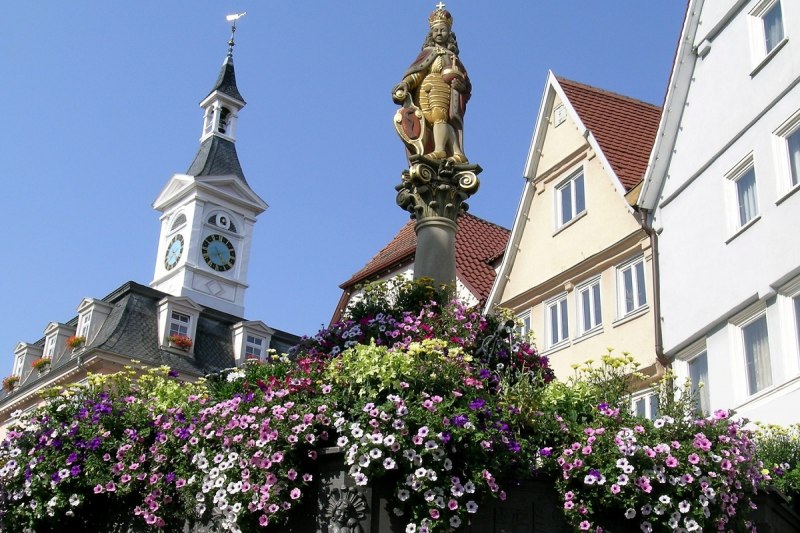 Der blumengeschmückte Brunnen am Aalener Marktplatz mit der Kirche, dem Spionrathaus und Fachwerkhäusern im Hintergrund